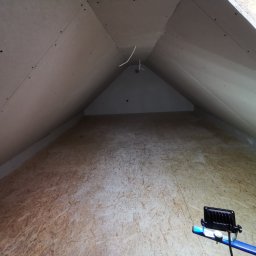 Iberhome - Poddasze z płytami karton-gips na skosach i podłodze z płyt OSB, oświetlone lampą roboczą. Widoczne przewody elektryczne.