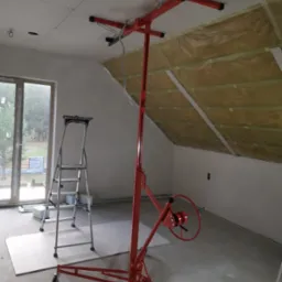 Montaż płyty G-K na poddaszu z użyciem podnośnika. Widoczna izolacja i drabina. Prace wykończeniowe w trakcie realizacji, surowe wnętrze.