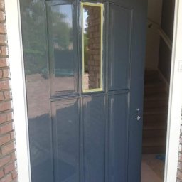 Iberhome - Świeżo pomalowane na szaro drzwi wejściowe z taśmą malarską na szybie, widok na schody wewnątrz domu. Ceglana elewacja z białym obramowaniem.