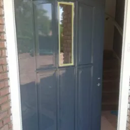 Świeżo pomalowane na szaro drzwi wejściowe z taśmą malarską na szybie, widok na schody wewnątrz domu. Ceglana elewacja z białym obramowaniem.