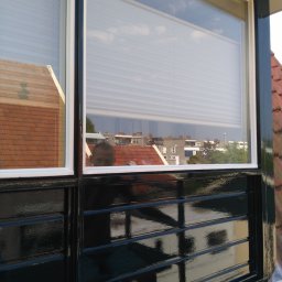 Iberhome - Błyszczące, świeżo pomalowane na czarno okno z białymi ramami. W szybie odbija się budynek i niebo. Na dole folia zabezpieczająca.