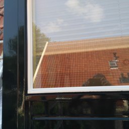 Iberhome - Częściowy widok na odnowione, czarne obramowanie okna z białymi listwami, z drabiną opartą o dach w tle. W szybie odbicie dachu i nieba.