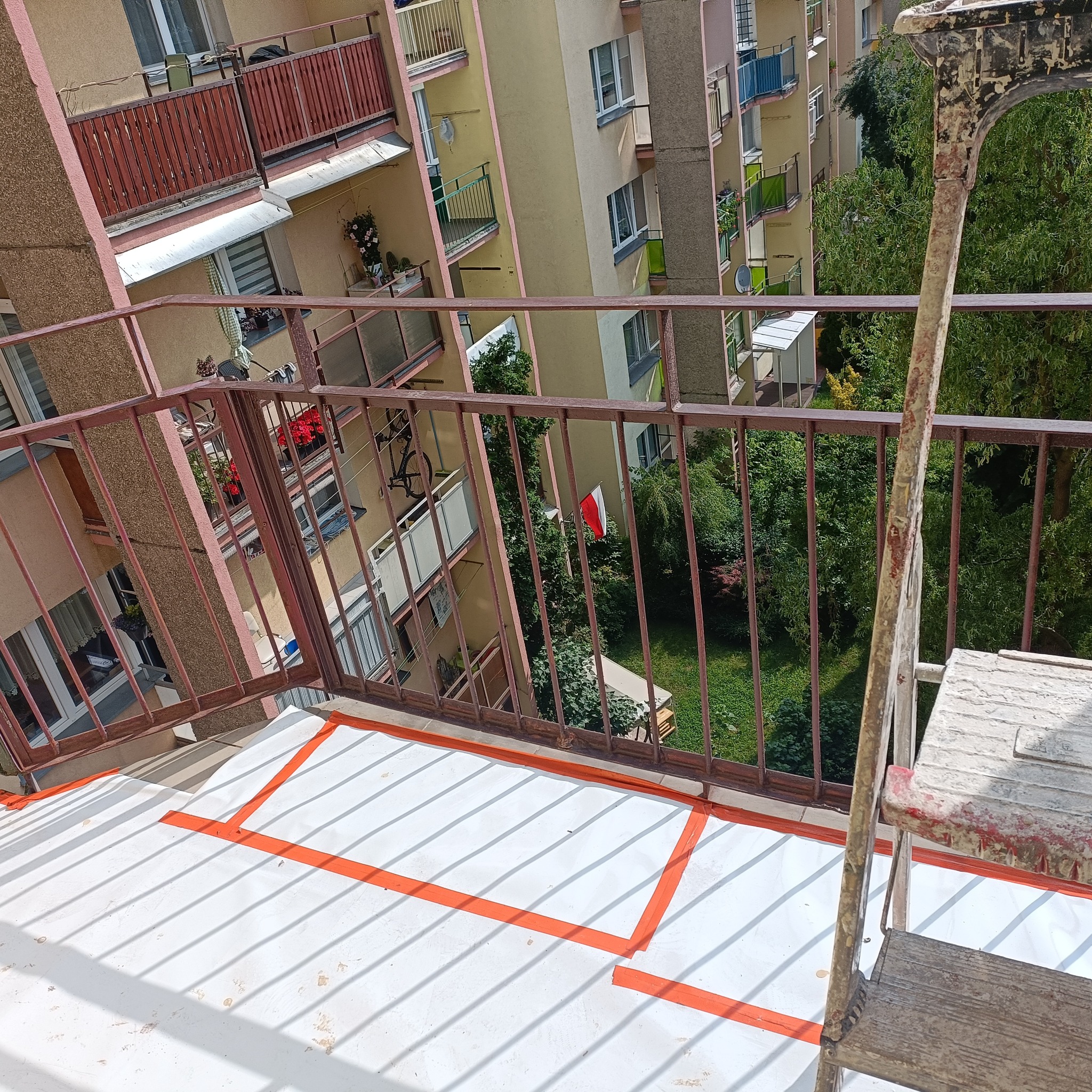 Balkon zabezpieczony folią i taśmą malarską przed malowaniem. W tle bloki mieszkalne i zieleń. Widoczna drabina malarska.