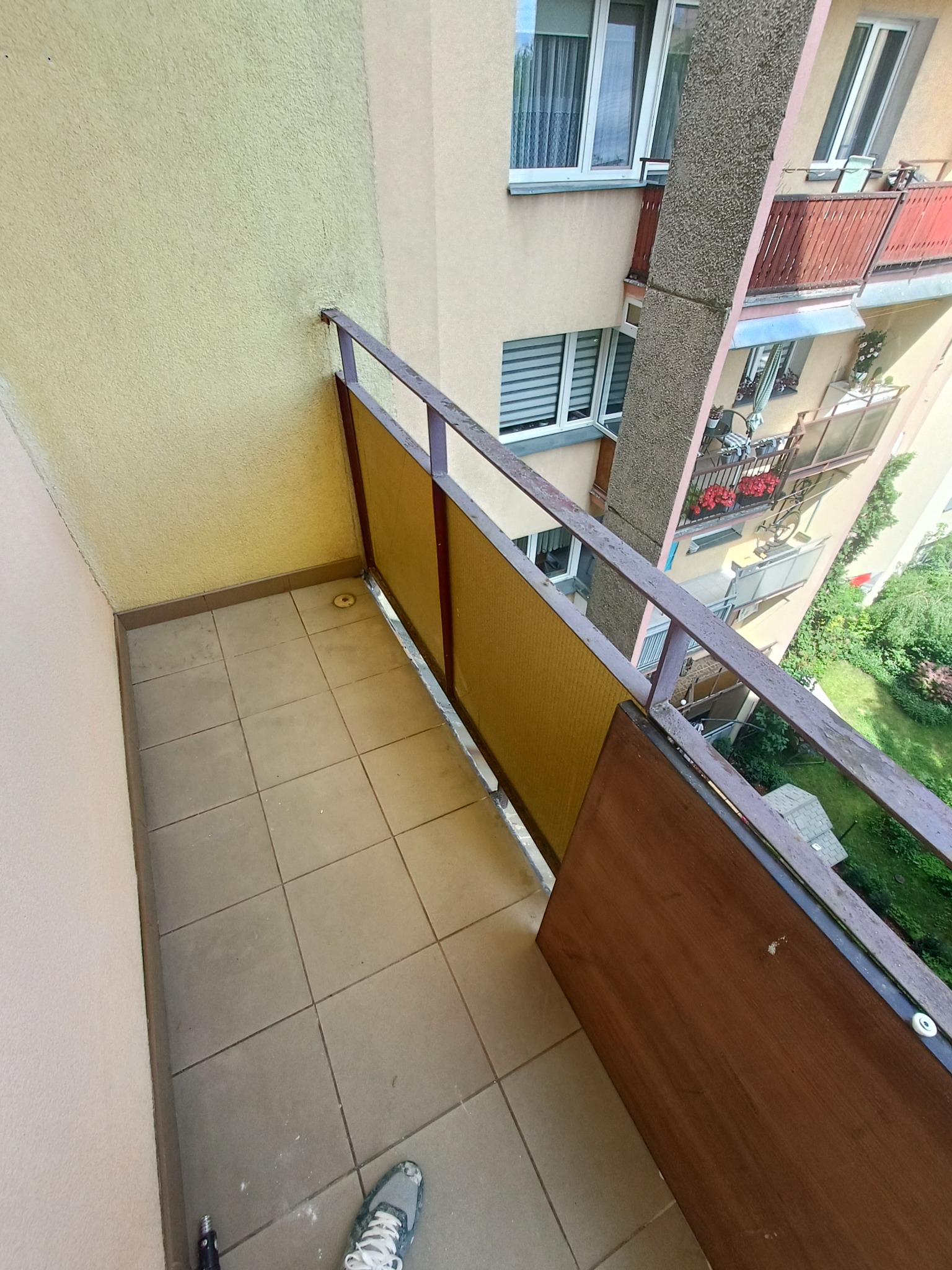Balkon z brązowymi płytkami podłogowymi w Kaliszu, widok z góry. Na podłodze leży but. Widoczne barierki i fragmenty sąsiednich balkonów.