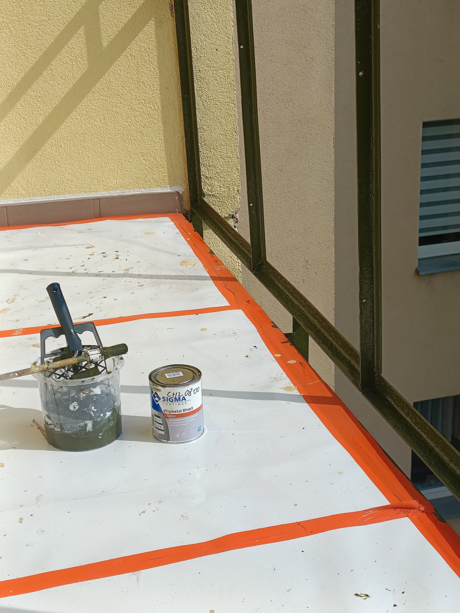 Malowanie balustrady balkonowej farbą Sigma, widoczny pędzel, wiadro z farbą i zabezpieczona folią podłoga. Świeżo pomalowane elementy na pomarańczowo.