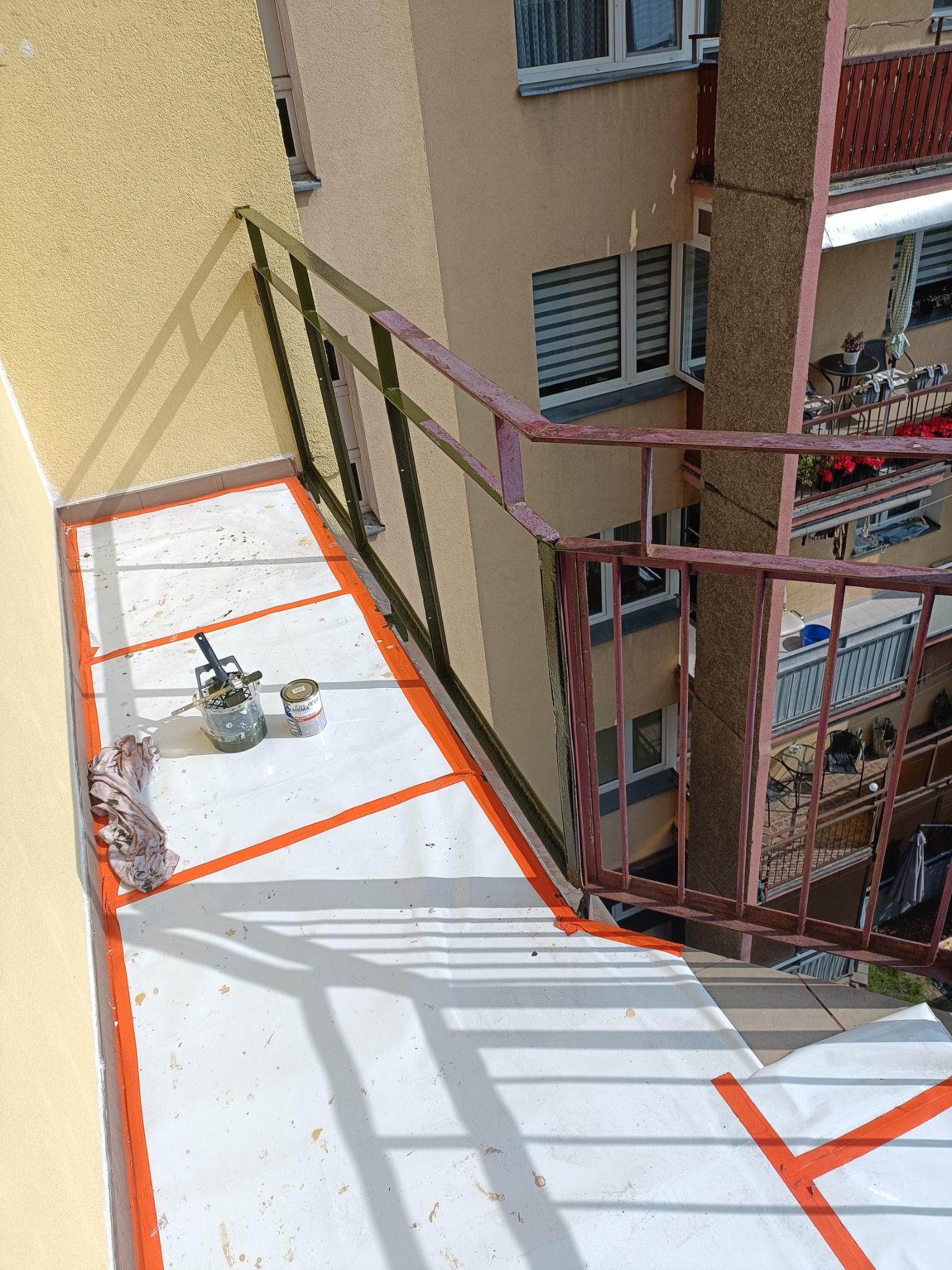 Biała podłoga balkonu zabezpieczona taśmą malarską, obok puszka farby i wałek. Widok z góry na balkon w bloku w trakcie malowania. Balustrada z metalowych prętów.
