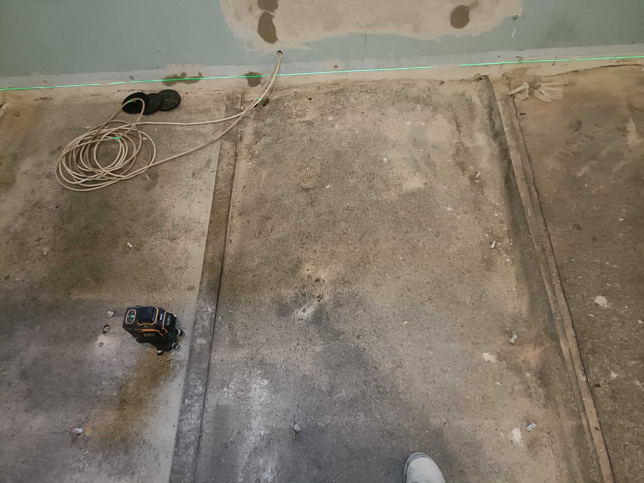 Przygotowanie podłoża pod gładzie: Laser poziomujący, kable, i fragment stopy w szarym obuwiu na surowej podłodze. Widoczny fragment sufitu z płytami G-K.