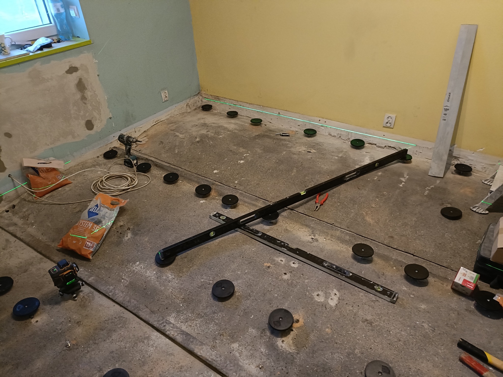 Przygotowanie podłoża pod wylewkę samopoziomującą: laser krzyżowy, poziomnice i regulowane podkładki dystansowe na surowym betonie w remontowanym pomieszczeniu.