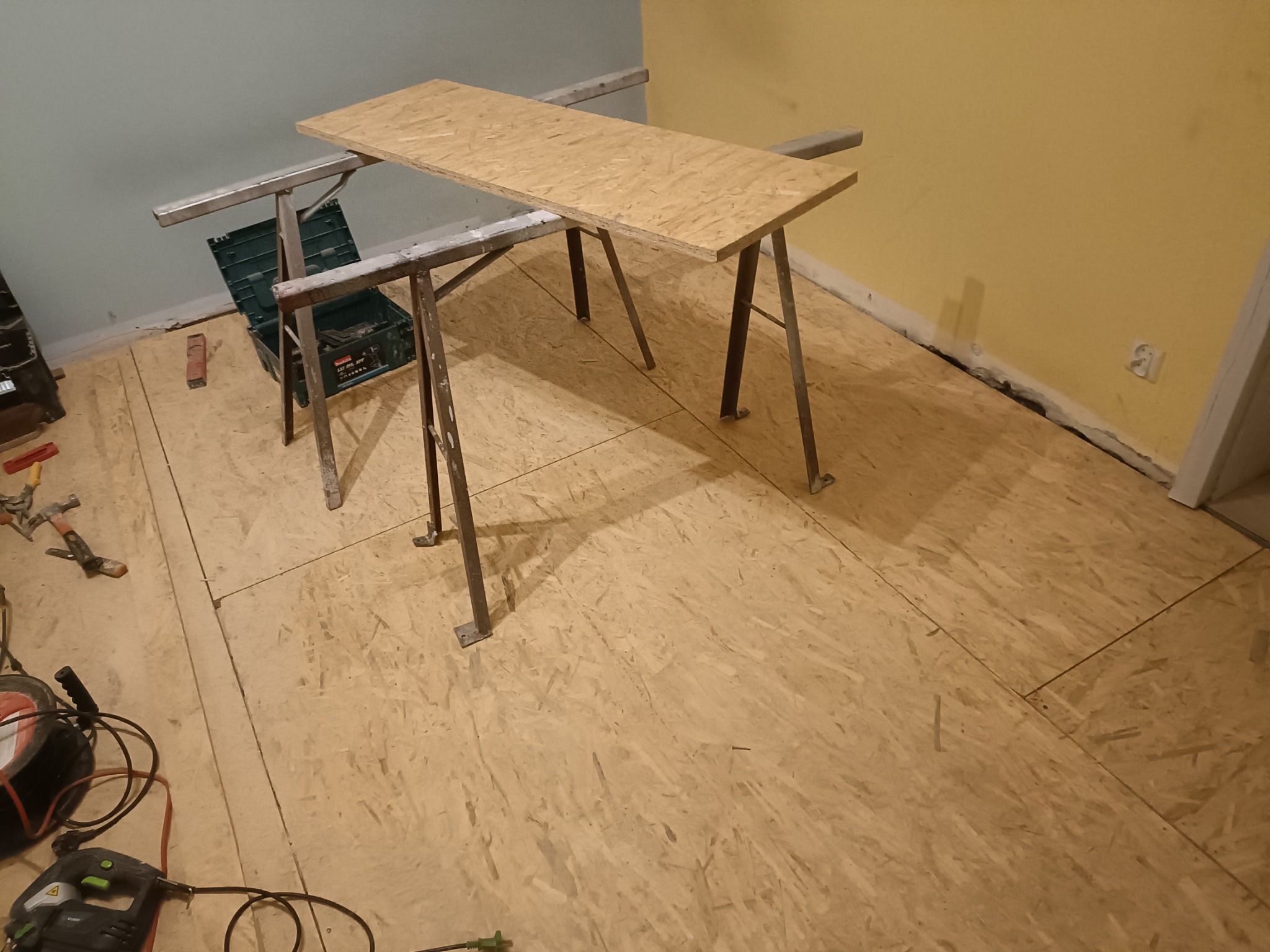 Płyta OSB ułożona na podłodze, widoczne narzędzia i skrzynka narzędziowa. Przygotowanie do prac wykończeniowych w pomieszczeniu z żółtą i niebieską ścianą.