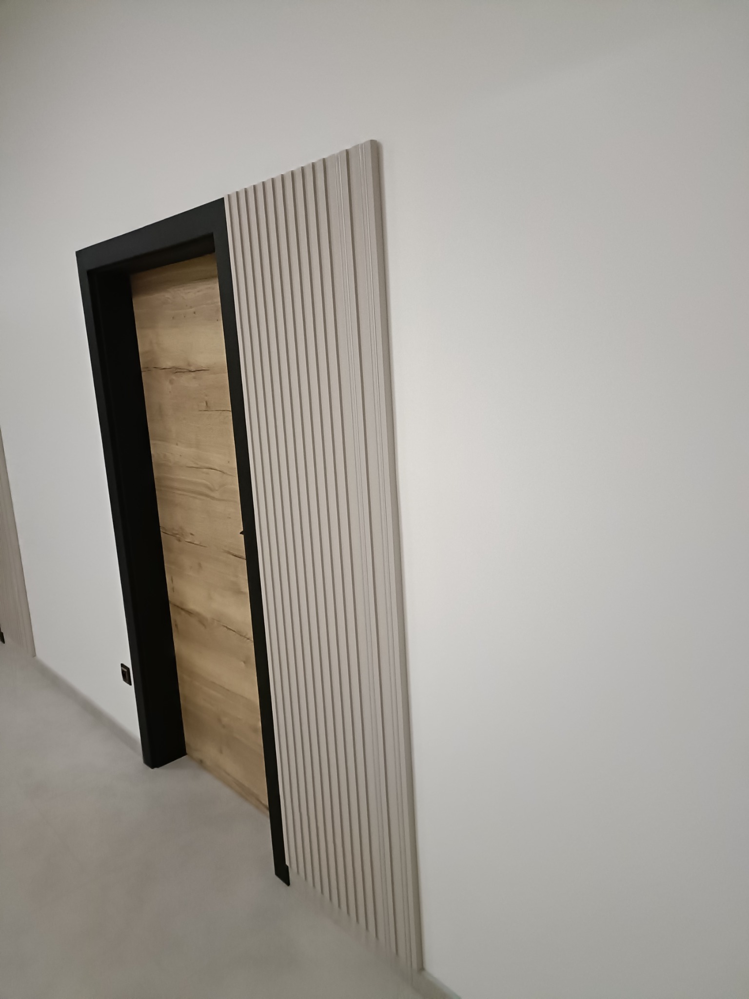 Nowoczesne wykończenie wnętrza: drewniane drzwi z czarną ościeżnicą i dekoracyjną, pionową listwą na białej ścianie. Minimalistyczny design.