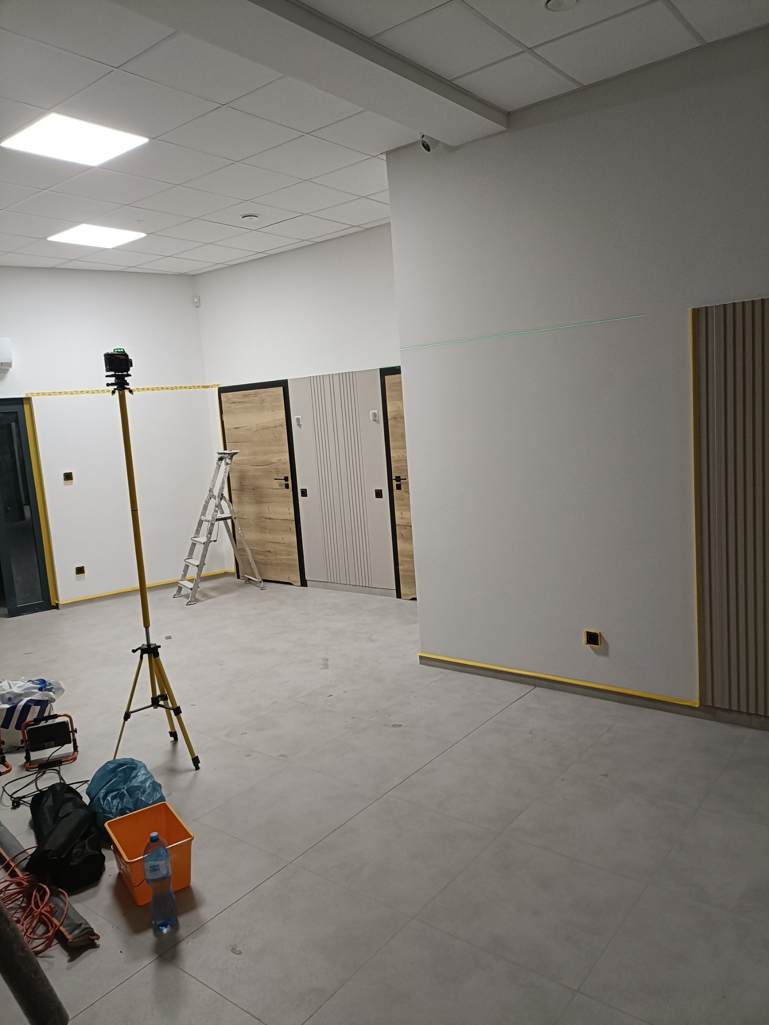 Wnętrze w trakcie prac wykończeniowych: laser krzyżowy na statywie, drabina, zabezpieczone taśmą ściany, drzwi i listwy dekoracyjne. Widok na sufit kasetonowy.