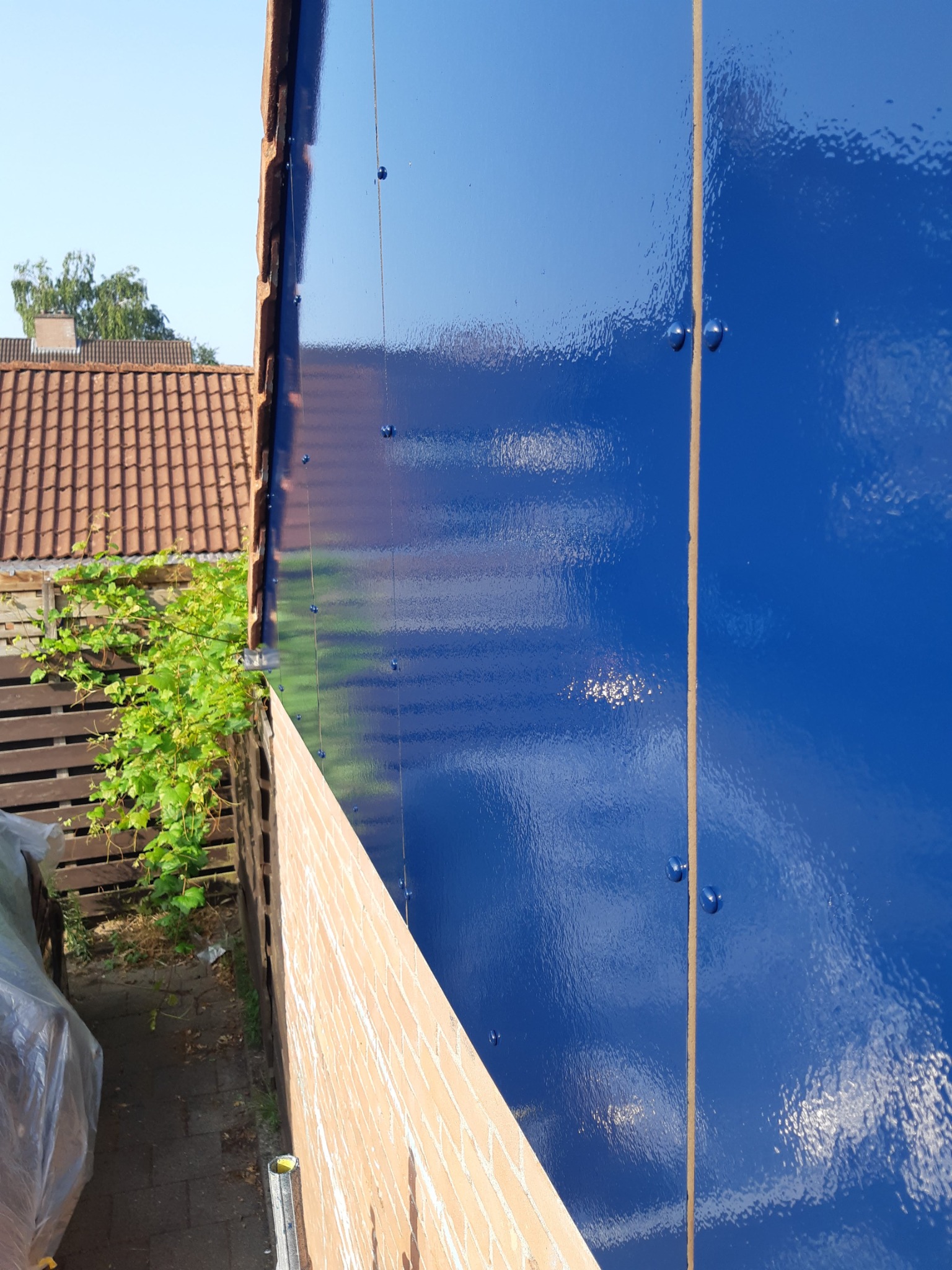 Niebieska, błyszcząca elewacja budynku z widocznymi śrubami mocującymi, sfotografowana z dołu, z fragmentem ceglanego muru i zieleni w tle.