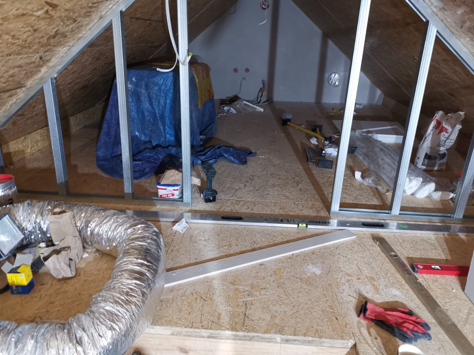 Montaż stelaża z profili metalowych na poddaszu, widoczne narzędzia, materiały izolacyjne i wentylacja. Prace wykończeniowe w trakcie realizacji.