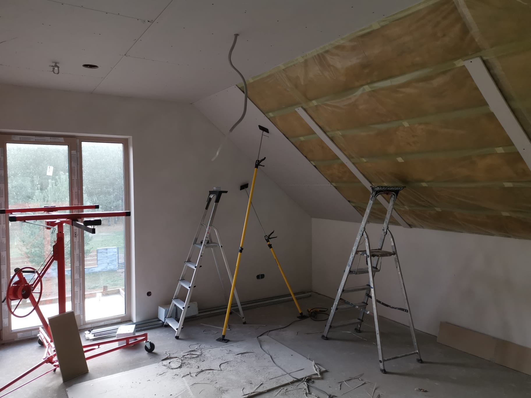 Poddasze w trakcie prac wykończeniowych: ocieplenie wełną, montaż płyt gipsowych. Widoczne drabiny, podnośnik do płyt i wystające przewody.