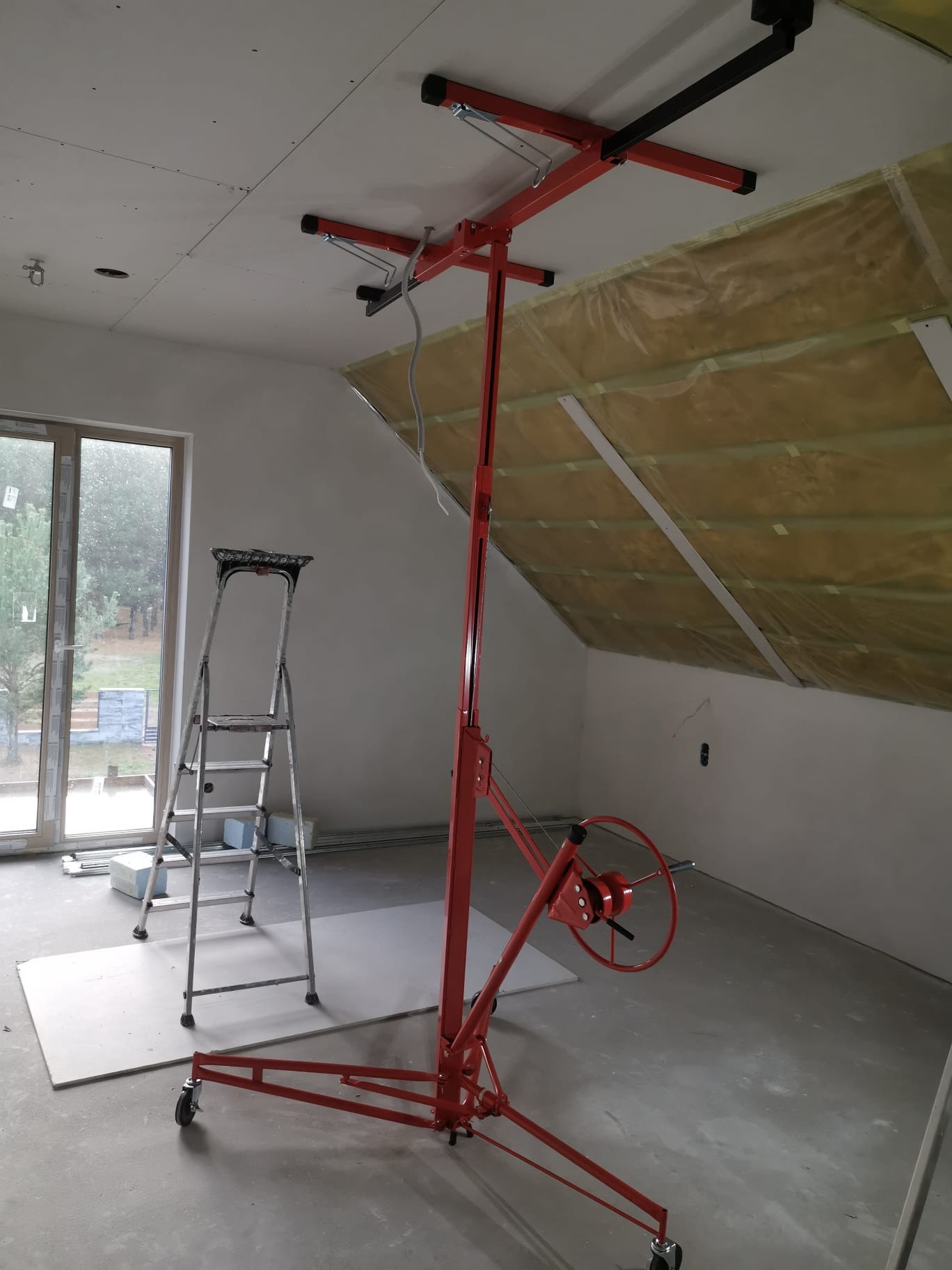 Montaż płyty G-K na poddaszu z użyciem podnośnika. Widoczna izolacja i drabina. Prace wykończeniowe w trakcie realizacji, surowe wnętrze.