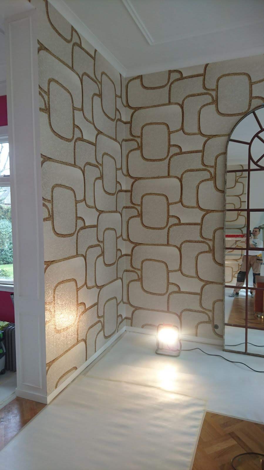 Wnętrze po remoncie z wzorzystą tapetą w stylu retro, zabezpieczona podłoga i widoczne odbicie osoby w lustrze. Nowoczesne wykończenie ścian.