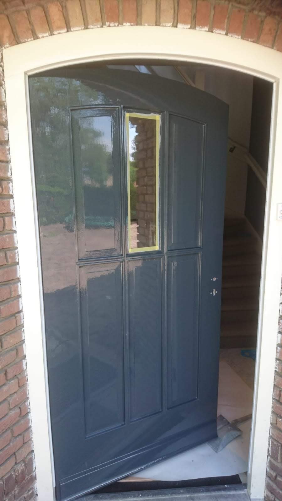 Świeżo pomalowane na szaro drzwi wejściowe z taśmą malarską na szybie, widok na schody wewnątrz domu. Ceglana elewacja z białym obramowaniem.