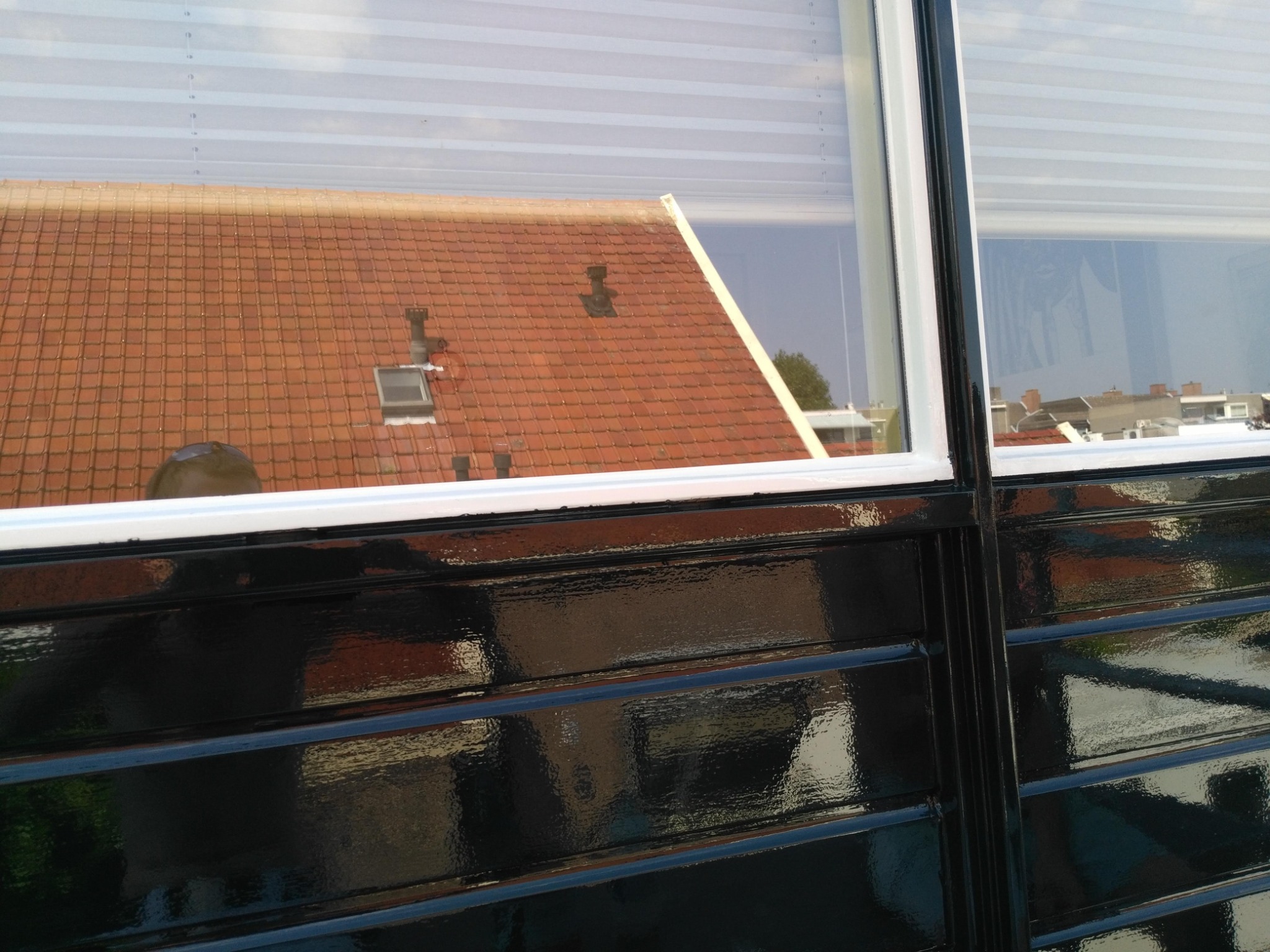 Błyszcząca, czarna, świeżo pomalowana boazeria okienna odbija czerwoną dachówkę budynku zza okna z żaluzjami. Widoczne detale okien i refleksy światła.