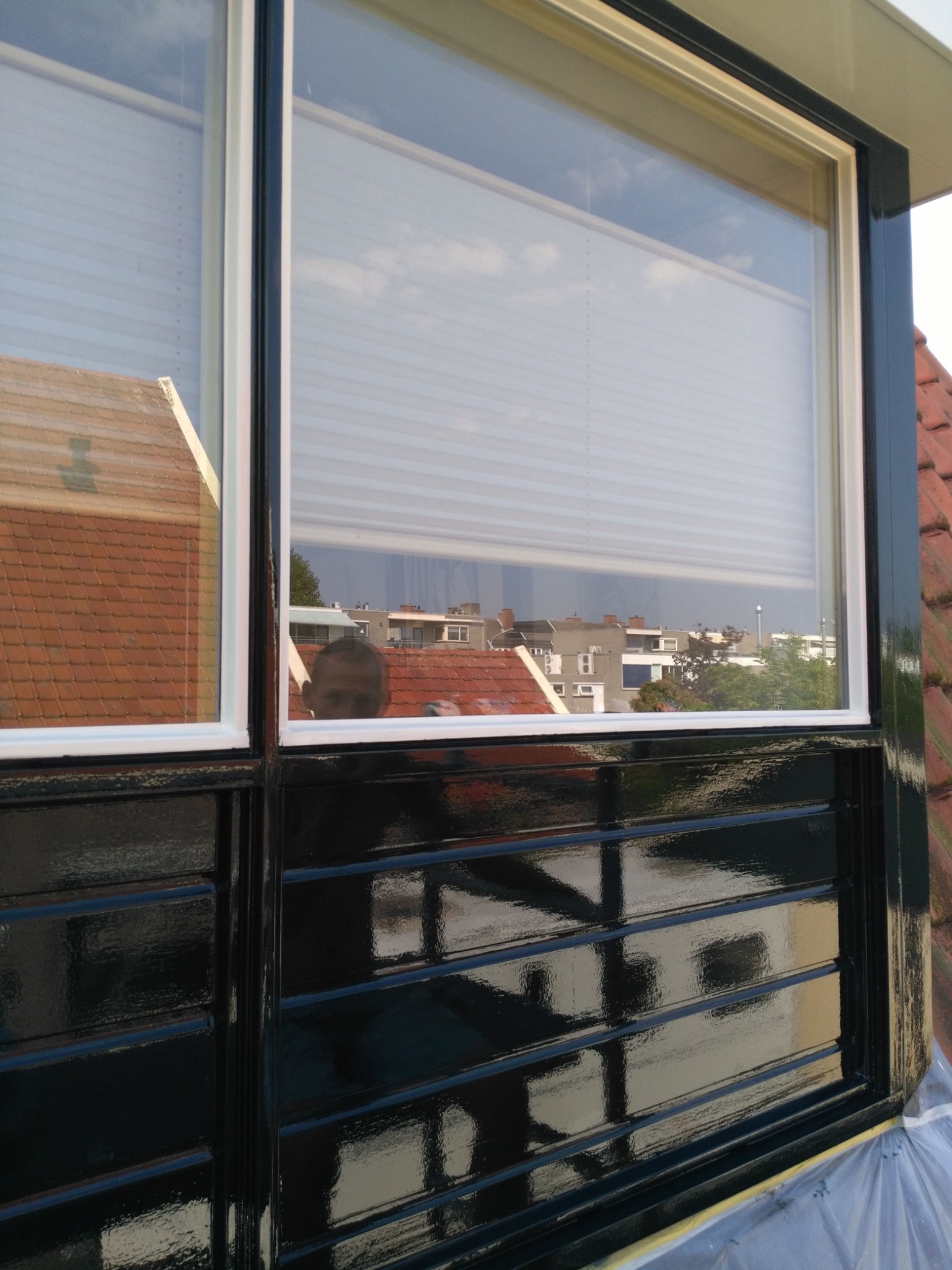 Błyszczące, świeżo pomalowane na czarno okno z białymi ramami. W szybie odbija się budynek i niebo. Na dole folia zabezpieczająca.