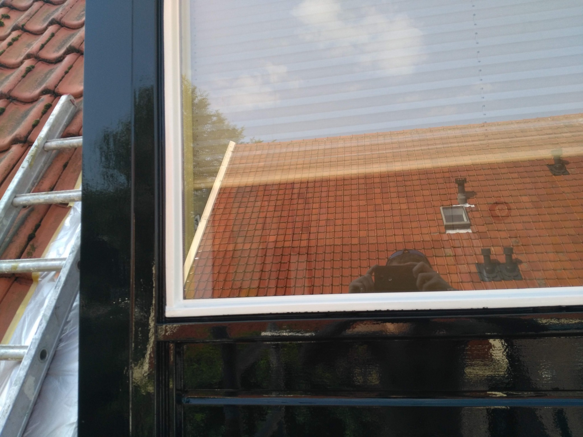 Częściowy widok na odnowione, czarne obramowanie okna z białymi listwami, z drabiną opartą o dach w tle. W szybie odbicie dachu i nieba.