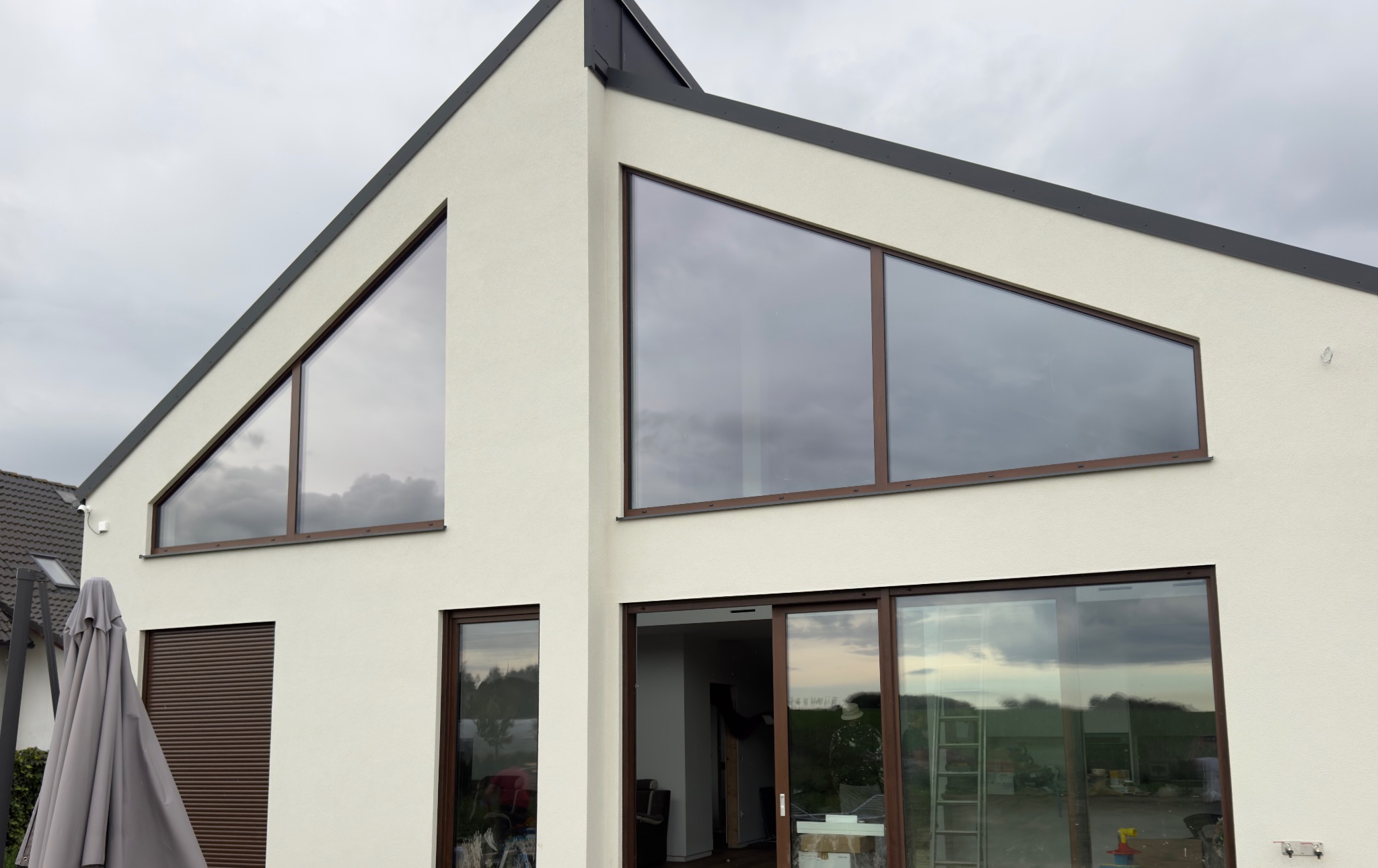 Nowoczesny dom z jasną elewacją i dużymi oknami o nietypowych kształtach, brązowe ramy okienne, widoczne odbicie nieba w szybach.