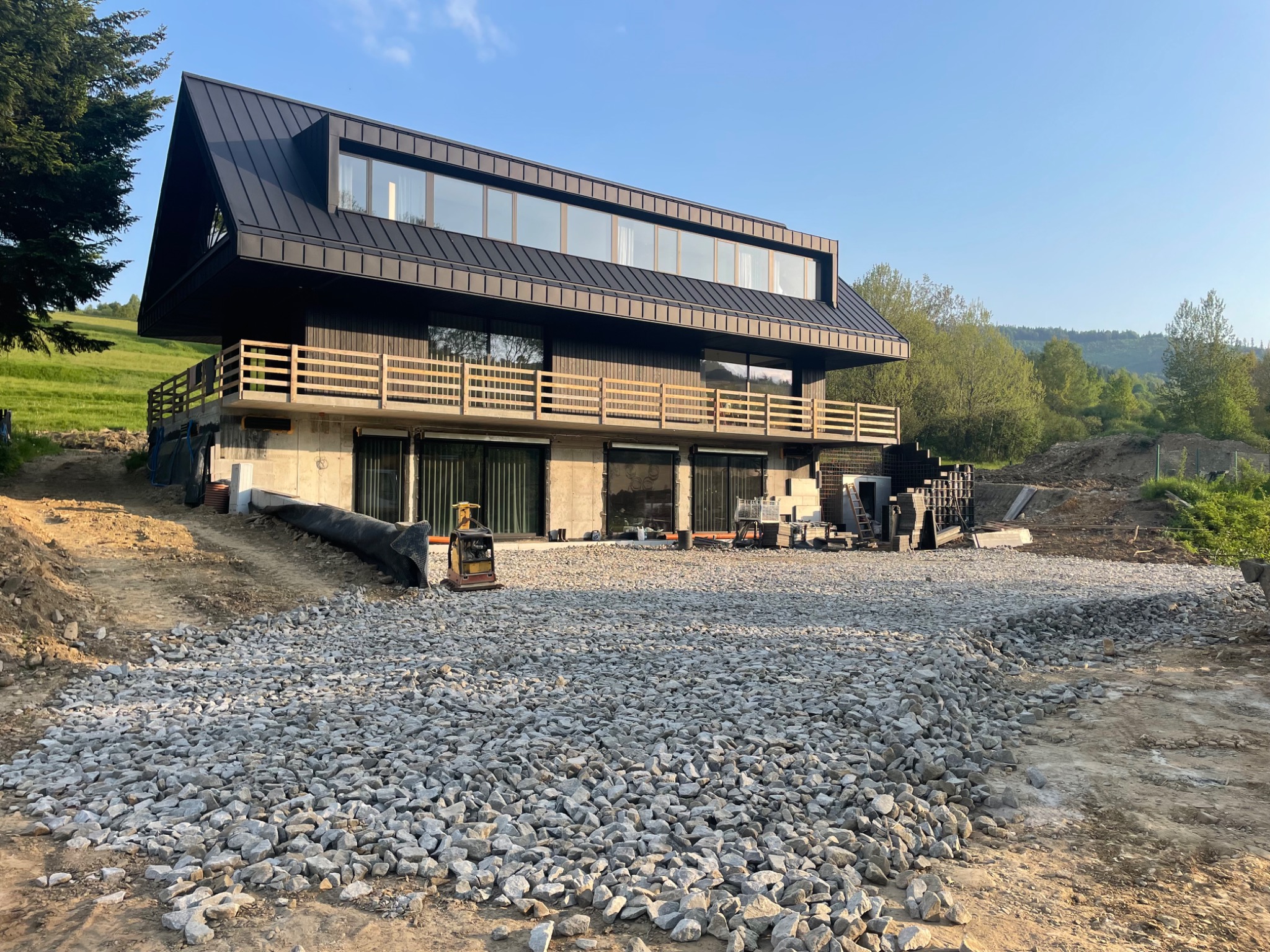 Nowoczesny dom z czarnym dachem i drewnianą balustradą, teren przygotowany pod dalsze prace, z widocznymi kamieniami i mini-koparką.