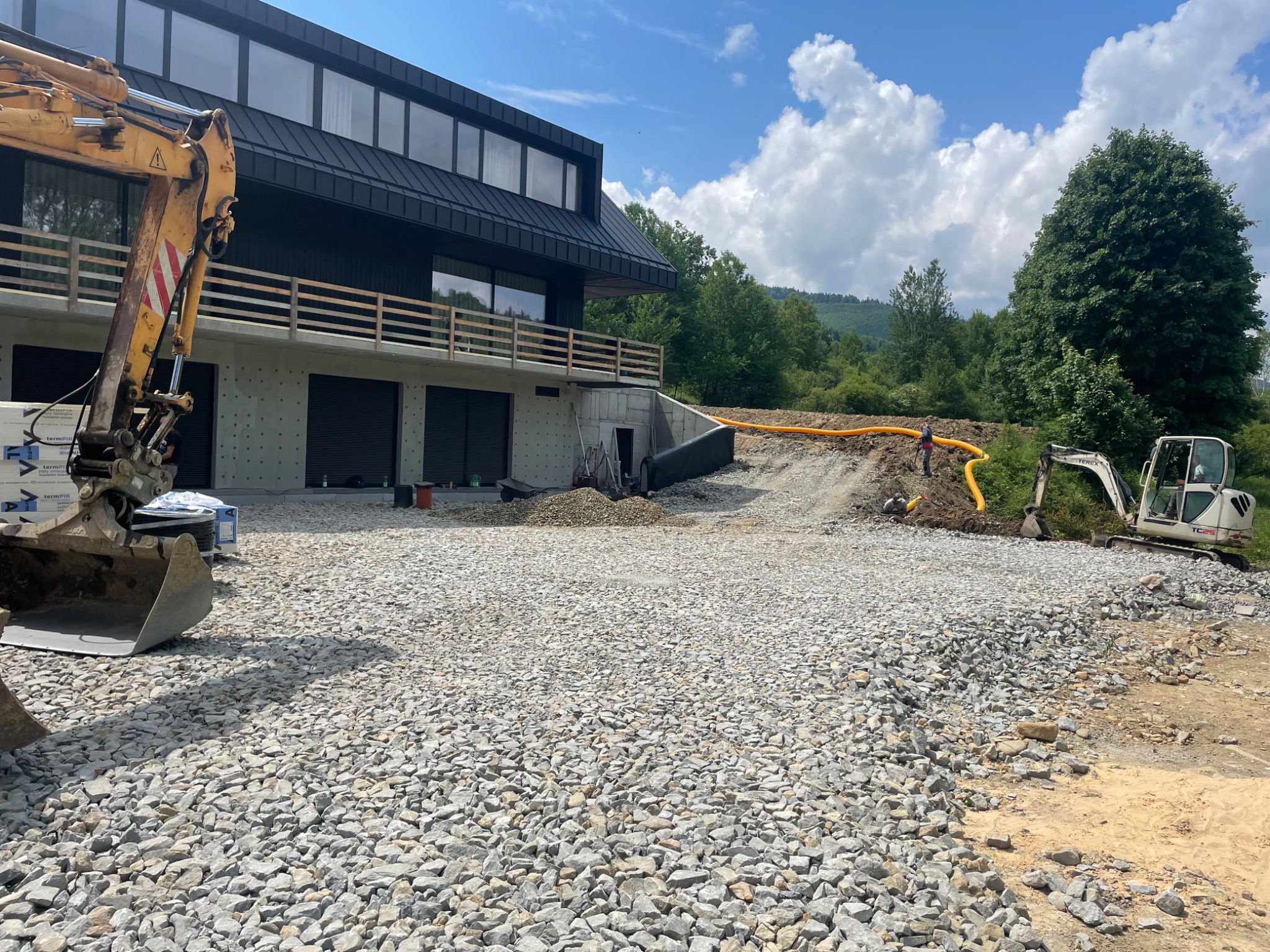 Żwirowe podłoże przed nowoczesnym domem z dwoma koparkami w tle i żółtymi rurami na wzniesieniu.