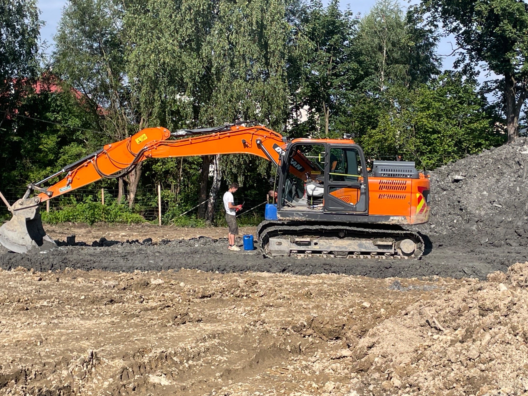 Pomarańczowa koparka gąsienicowa Doosan wyrównuje teren, obok stoi mężczyzna z tabletem, w tle drzewa i hałda ziemi.