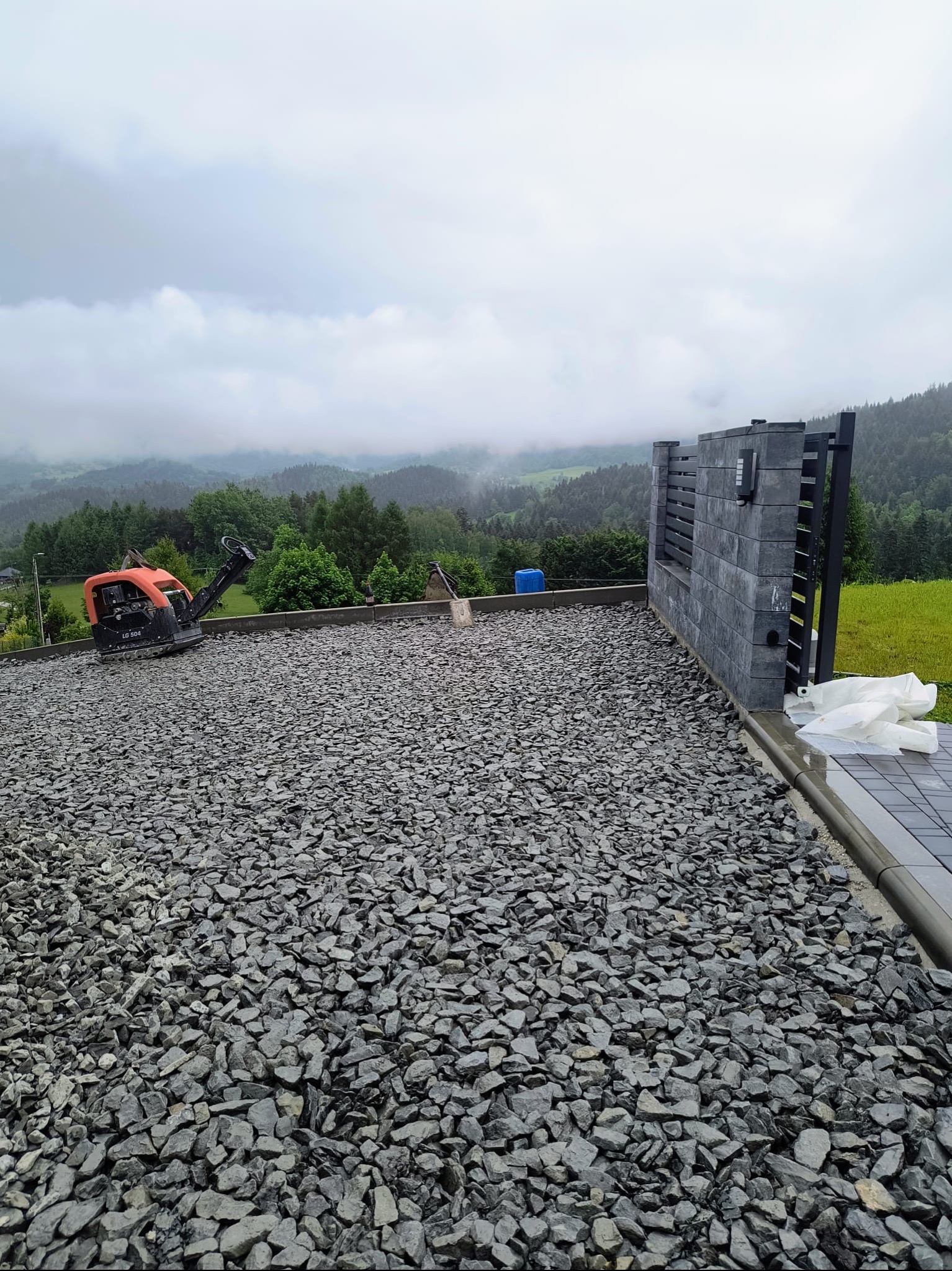 Przygotowanie terenu pod budowę: zagęszczarka wibrująca na warstwie tłucznia, widok na wzgórza i nowoczesne ogrodzenie z pustaków betonowych.