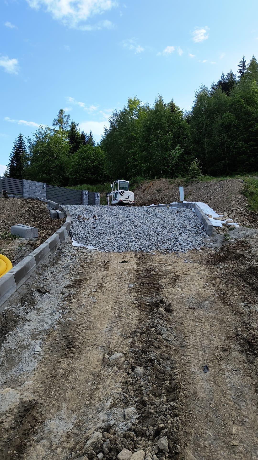 Przygotowanie podłoża pod drogę dojazdową: ubity grunt z widocznymi śladami opon, warstwa gruzu, obrzeża betonowe i walec drogowy w tle, teren pagórkowaty z drzewami.