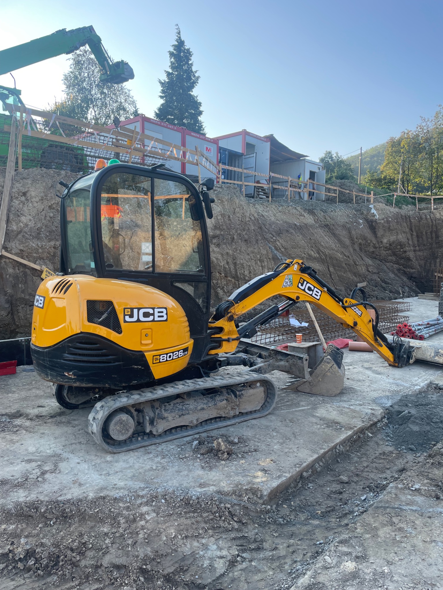 Żółta mini koparka JCB 8026 CTS na betonowej płycie fundamentowej, w tle wykop ziemny i kontenery budowlane