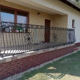 Kuta, czarna balustrada balkonowa z ozdobnymi rozetami na tle elewacji domu z cegły i piaskowca, widok z poziomu trawnika.