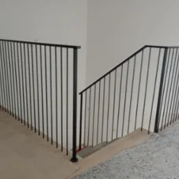 Minimalistyczna, czarna balustrada metalowa na schodach w surowym wnętrzu, widoczne zabezpieczenia podłogi, prosta forma poręczy.