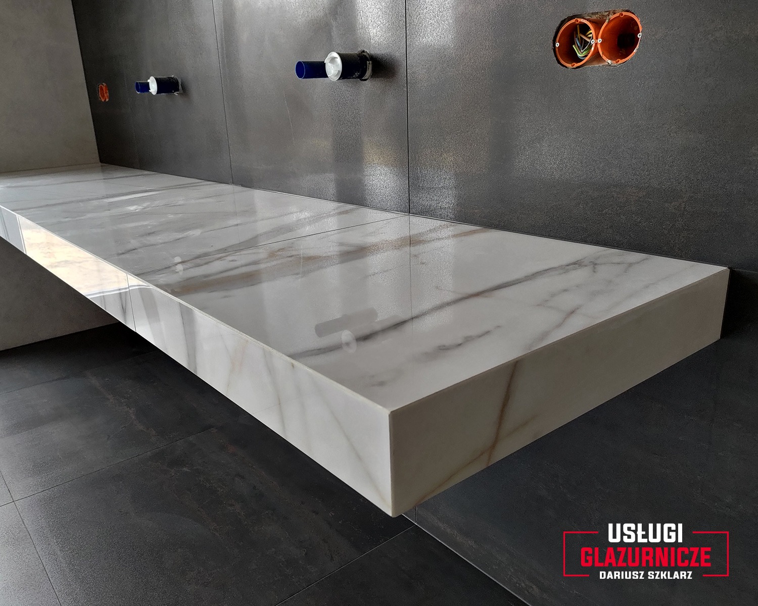 Łazienka w Kolbuszowej z wykończonym blatem z płytek imitujących marmur, dopasowanych do ciemnych płytek podłogowych i szarej ściany, widoczne instalacje wodne i elektryczne.