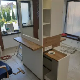 Biała zabudowa meblowa z drewnianymi akcentami i lustrem, oświetlona od góry, z otwartą szufladą i widocznymi gniazdkami elektrycznymi w pomieszczeniu z oknem i krzesłem fryzjerskim w tle.