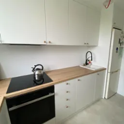 Jasna kuchnia z białymi frontami szafek z okrągłymi, złotymi uchwytami, drewnianym blatem, czarną baterią i płytą indukcyjną z czajnikiem.