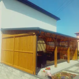 Drewniana konstrukcja z zasuwanymi panelami, prawdopodobnie altana lub pergola, przylegająca do białego budynku z ciemnym dachem, z widocznymi kwiatami w donicach i narzędziami na pierwszym planie.