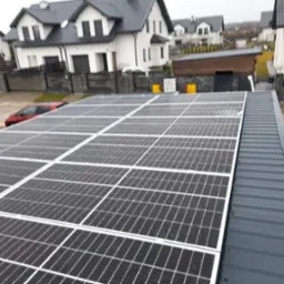 Panel fotowoltaiczny na dachu budynku mieszkalnego w pochmurny dzień, widoczne krople wody na panelach i tło z domami jednorodzinnymi.