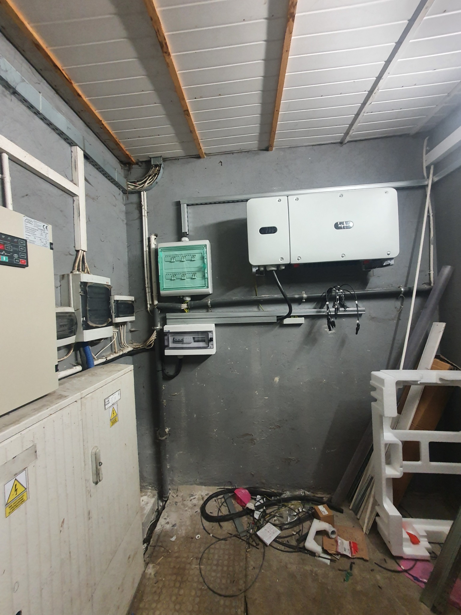 Wnętrze pomieszczenia z zamontowanym inwerterem fotowoltaicznym, rozdzielnicą elektryczną i przewodami, widoczne elementy konstrukcji sufitu i ściany pomalowanej na szaro, na podłodze leżą kable...