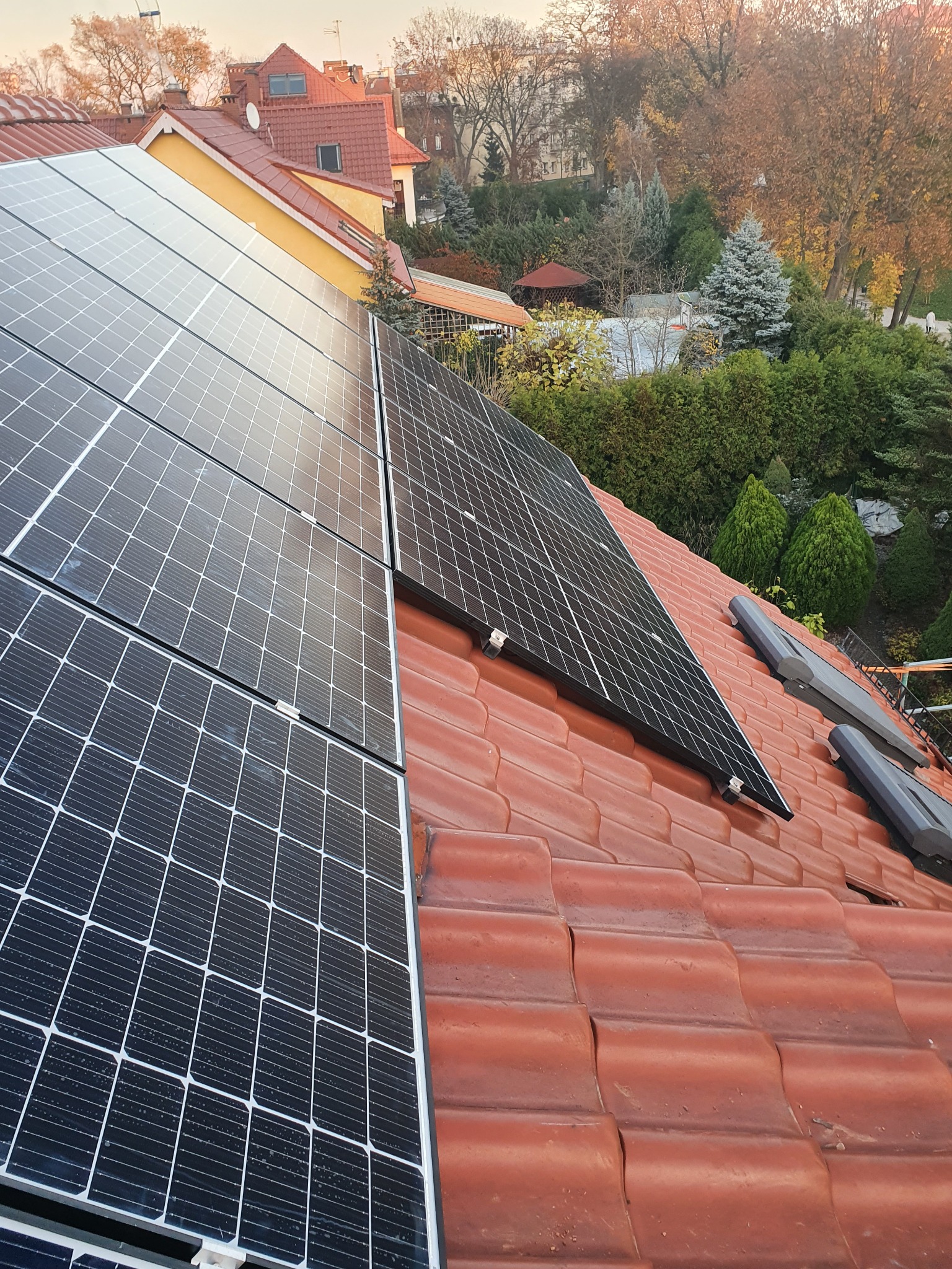 Panele fotowoltaiczne zamontowane na czerwonej dachówce, widok z góry na tle zieleni i budynków w oddali.