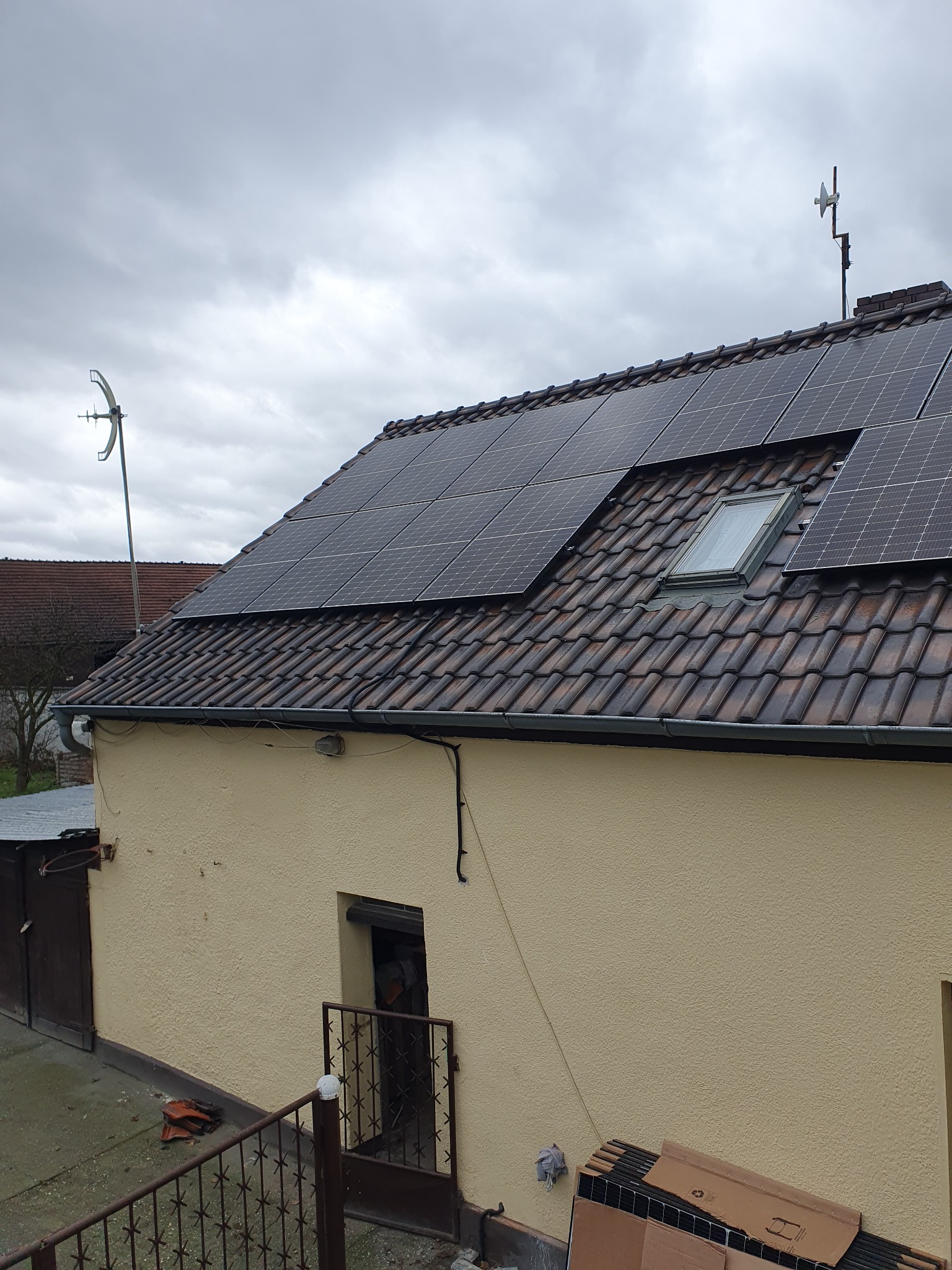 Panele słoneczne na dachu budynku z brązową dachówką, widoczne okno dachowe i dwie anteny, pochmurne niebo.