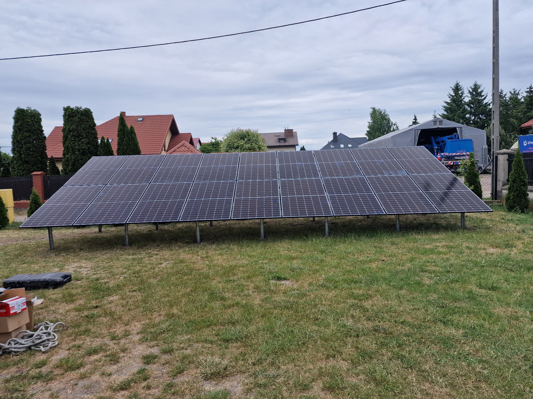 Rząd paneli fotowoltaicznych na metalowych nogach, ustawionych na trawniku przed domami jednorodzinnymi, z widocznymi akcesoriami montażowymi obok.