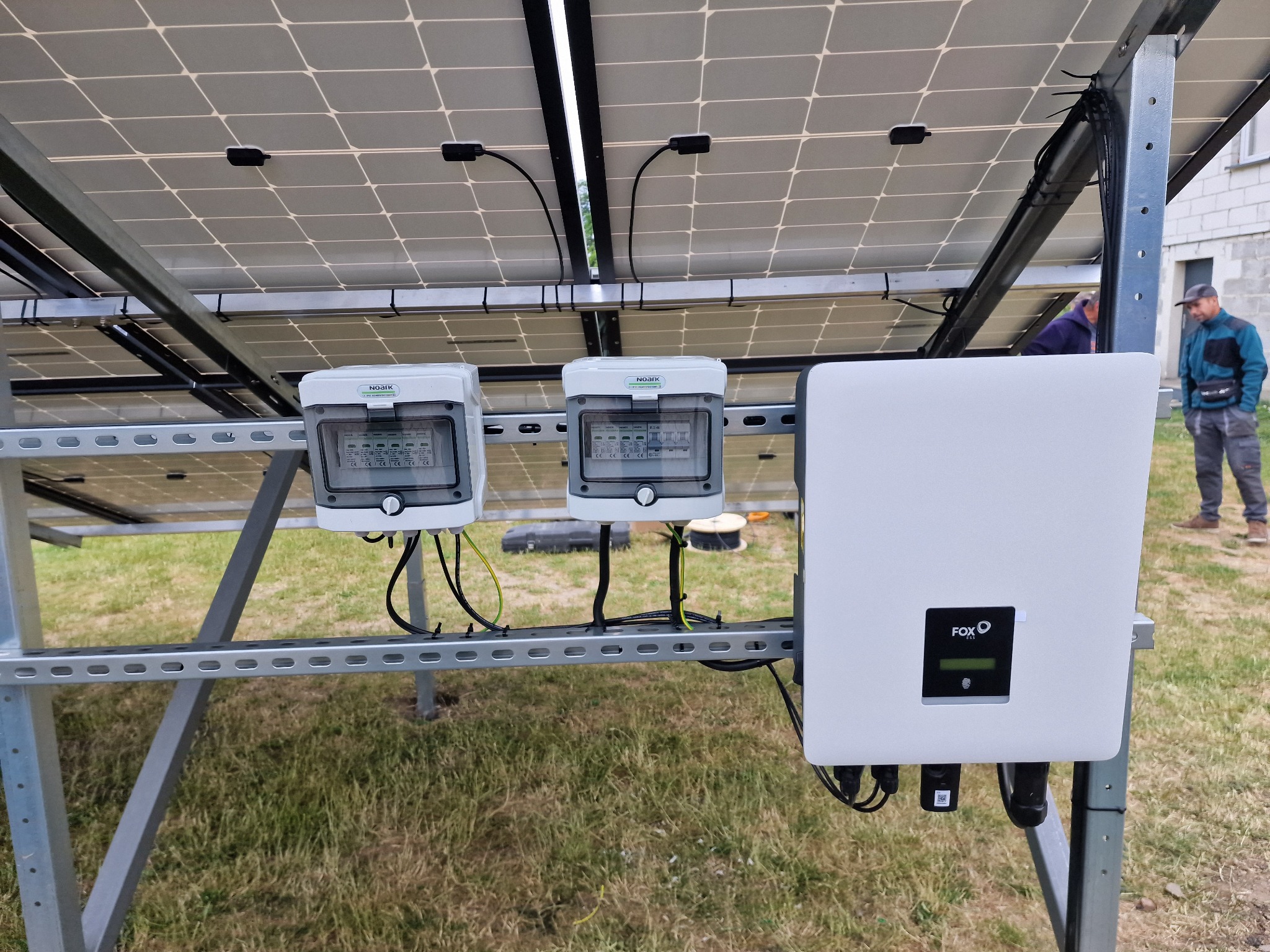 Dwa białe rozdzielacze NOARK i biały falownik FOX ESS zamontowane na metalowej konstrukcji pod panelami słonecznymi, widoczne kable i trawa w tle, dwóch mężczyzn w oddali.