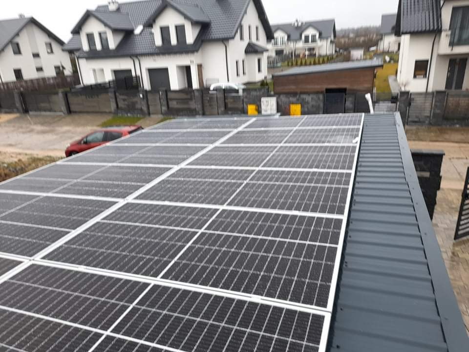 Panel fotowoltaiczny na dachu budynku mieszkalnego w pochmurny dzień, widoczne krople wody na panelach i tło z domami jednorodzinnymi.