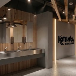 PP Paula Tylek - Nowoczesna toaleta publiczna z drewnianymi akcentami, lustrami i metalowymi umywalkami. Widoczny znak 'Kotelnica Białczańska' i piktogram kobiety.