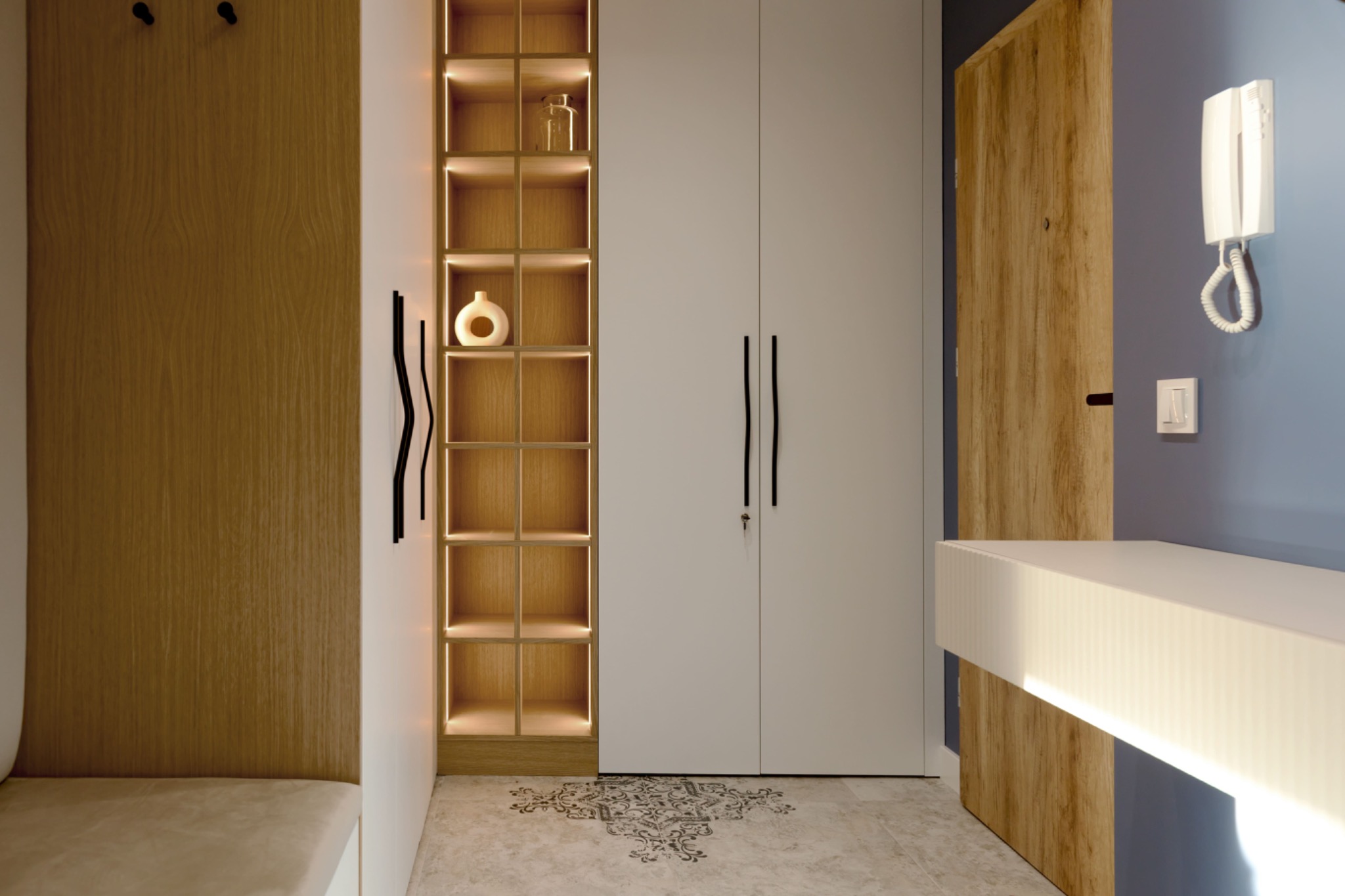 Nowoczesne wnętrze przedpokoju z szafą w zabudowie, podświetlanymi półkami z dekoracjami, drewnianą boazerią i wzorzystą posadzką.