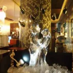Lodowa rzeźba pary małp na blacie baru, oświetlona od dołu, z dekoracyjną kolumną w tle i butelkami wiszącymi nad głową, prawdopodobnie element dekoracyjny imprezy.