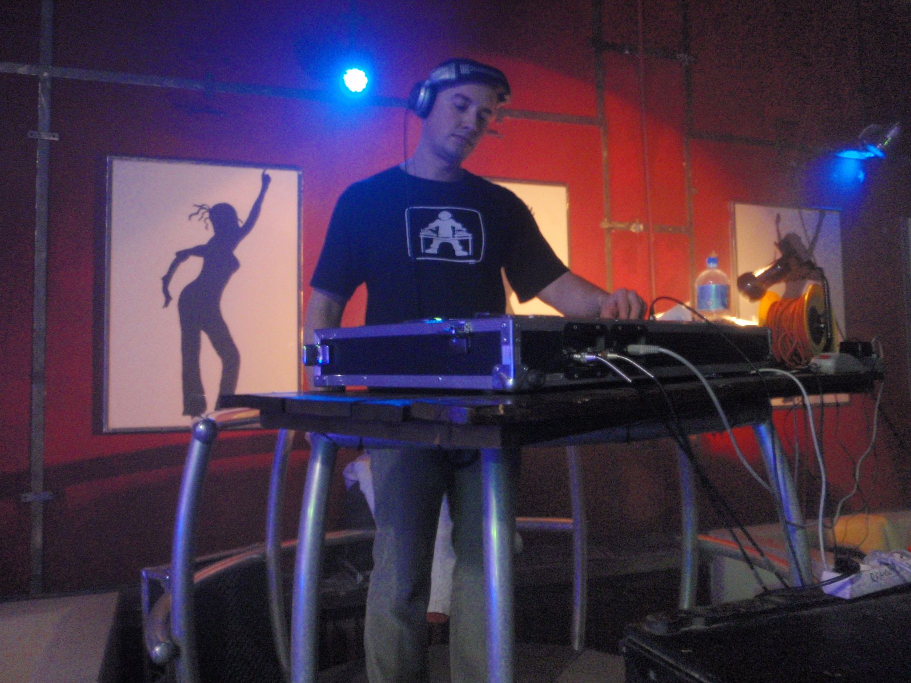 DJ w słuchawkach, miksujący muzykę na sprzęcie, w tle czerwona ściana z plakatami i niebieskie oświetlenie sceniczne.