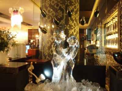 Lodowa rzeźba pary małp na blacie baru, oświetlona od dołu, z dekoracyjną kolumną w tle i butelkami wiszącymi nad głową, prawdopodobnie element dekoracyjny imprezy.