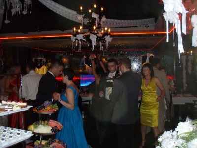 Uroczystość w ciemnym wnętrzu z dekoracjami, oświetleniem i bufetem; w tle tańczący ludzie, a w centrum para w strojach wieczorowych.